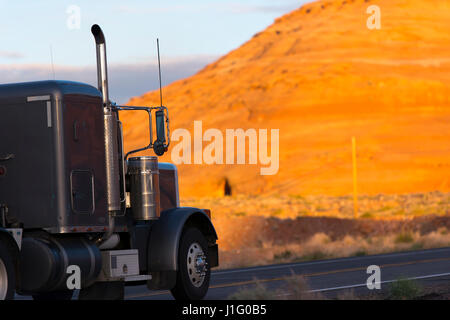 Dark classic big rig semi truck with long exhaust pipes passes by икшпре orange sandstone mountain in Page, Arizona, in hot summer day сarrying cargo Stock Photo