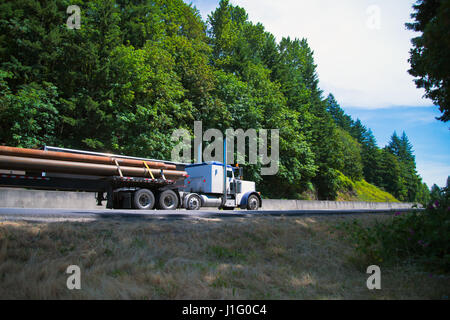 Classic white big rig semi truck with a long oversized load pipe on a flat bed trailer transports cargo on scenic highway surrounded by green trees Stock Photo
