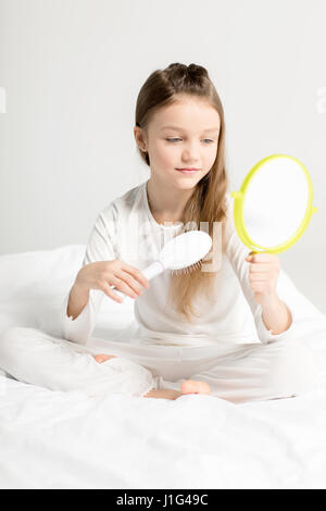 Adorable little girl in pajamas sitting on bed and combing hair Stock Photo