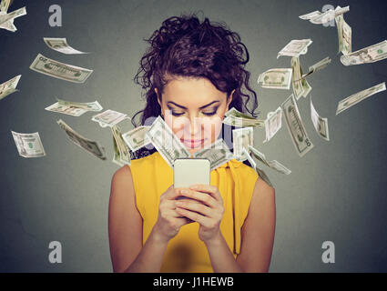 Online banking money transfer, e-commerce concept. Happy young woman using smartphone with dollar bills flying away from screen isolated on gray wall  Stock Photo
