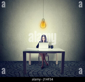 Young woman sitting at table working on laptop with bright light bulb above head Stock Photo