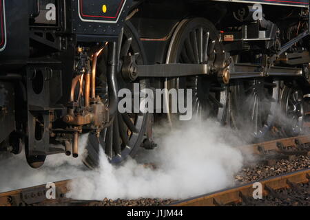 CLOSE UP LANDSCAPE VIEW OF THE WHEELS OF A STEAM LOCOMOTIVE IN MOTION WITH MUCH WHITE STEAM COMING FROM THE WHEELS AND ONTO THE TRACK Stock Photo