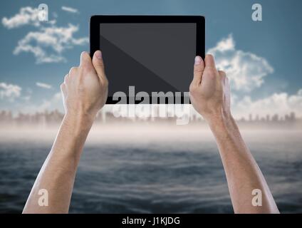 Digital composite of Hand with tablet against blurry skyline with water Stock Photo