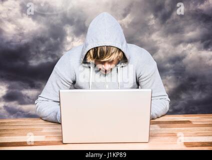 Digital composite of Criminal in hood on laptop in front of clouds Stock Photo