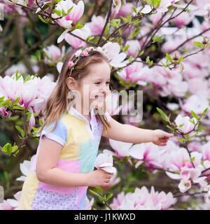 Happy little girl in pastel color dress and flower crown playing under pink magnolia tree picking big flowers in blooming spring garden. Child with ma Stock Photo