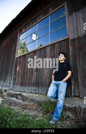 An asian student carrying a backpack leaning against an old barn Stock Photo
