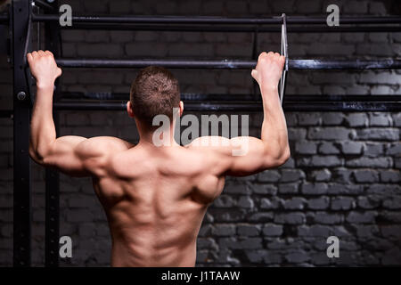 Rear view photo of muscular male doing exercises on horizontal bar against brick wall at the cross fit gym. Body and muscle. Power and energy. Healthy Stock Photo