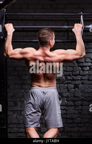 Rear view photo of muscular male doing exercises on horizontal bar against brick wall at the cross fit gym. Sportsman in the grey sportive shorts. Bod Stock Photo