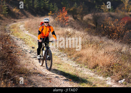 Cyclist in the orange jacket riding a bike on countryside road at the sunrise. Sportsman in the sportwear, white helmet, sunglasses and with red backp Stock Photo
