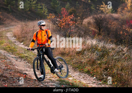 Cyclist in the orange jacket riding a bike on countryside road at the sunrise. Sportsman in the sportwear, white helmet, sunglasses and with red backp Stock Photo