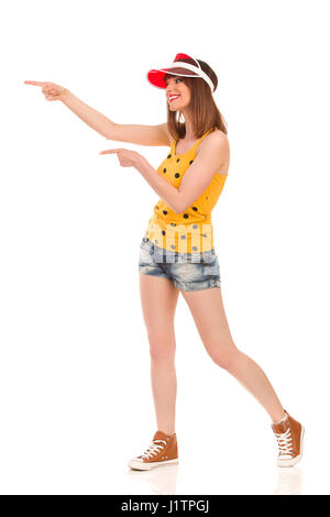 Young smiling woman in yellow dotted shirt, jeans shorts and brown sneakers is standing and pointing with both hands. Side view. Full length studio sh Stock Photo