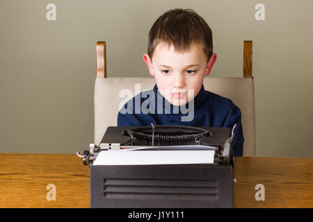 Cute little boy looks concentrated at work behind an old vintage black typewriter at home, thinking or reading his text Stock Photo