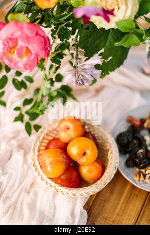 picnic, food, summer, holiday concept - close-up on the festively decorated table with beautiful pink peony and white rose flowers, fresh and dried fruits, nectarines, walnuts, prunes, tableware. Stock Photo