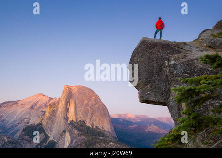 A fearless hiker is standing on an overhanging rock enjoying the view towards famous Half Dome at Glacier Point overlook at sunset, Yosemite NP, USA