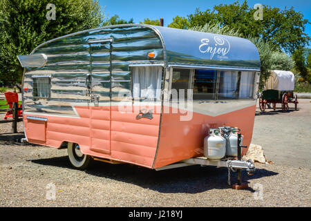 A salmon & silver colored classic, charming, vintage camper trailer in pristine condition awaits a tow, with an old covered wagon in background Stock Photo