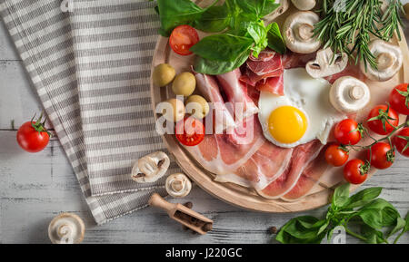 Healthy Mediterranean breakfast groceries: fried eggs, ham, vine tomatoes, mushrooms, basil, olives, rosemary on wooden tray. View from above Stock Photo