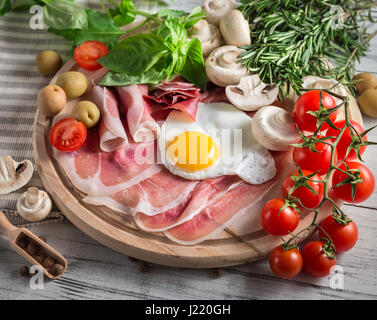 Healthy Mediterranean breakfast groceries: fried eggs, ham, vine tomatoes, mushrooms, basil, olives, rosemary on wooden tray Stock Photo