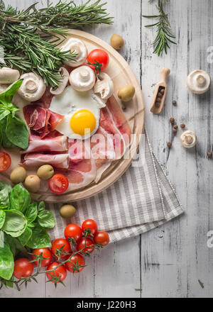 Healthy Mediterranean breakfast groceries: fried eggs, ham, vine tomatoes, mushrooms, basil, olives, rosemary on wooden tray. View from above Stock Photo