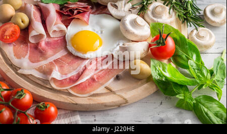 Healthy Mediterranean breakfast groceries: fried eggs, ham, vine tomatoes, mushrooms, basil, olives, rosemary on wooden tray Stock Photo