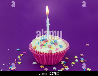 Bright purple background with single white cup cake in pink paper case and lit candle with colour star sprinkles for birthday celebration Stock Photo