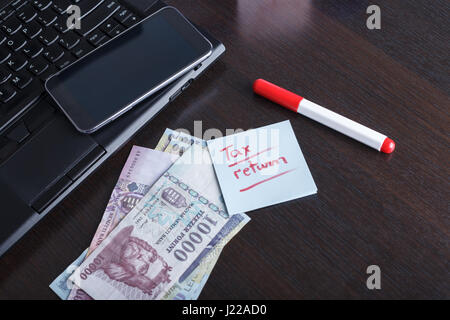 Handwritten note with tax return, refund text, formas and papers, notebook and laptop with mobile phone, cash on the table Stock Photo