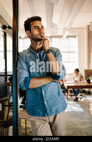 Portrait of young man standing at door looking away and thinking. Thoughtful young male at startup office with colleagues working in background. Stock Photo