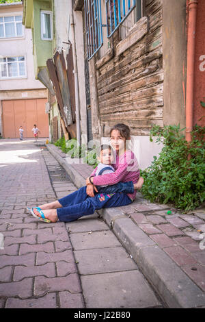 Girl sitting on the pavement in Balat area, behind the Grand Bazaar. Two girls, neatly dressed, older one holding a toddler next to old wooden house. Stock Photo