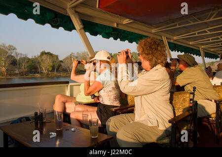 Women photographing animals on shore, on a sundowner cruise on the Zambezi River, Zimbabwe, Africa Stock Photo