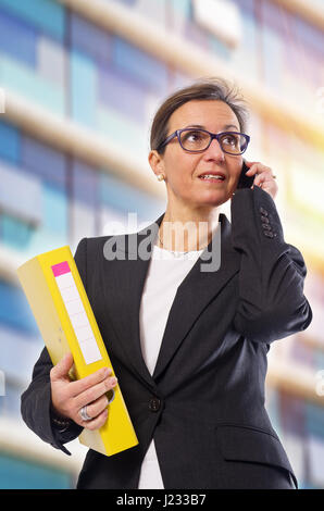 Brunette business woman with yellow folder and cellphone posing in front of office building Stock Photo