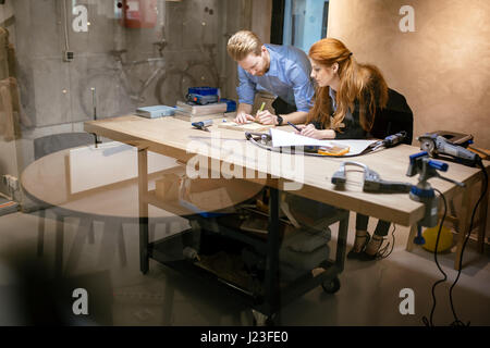 Creative designers working in workshop while standing at the desk full of tools and discussing ideas Stock Photo