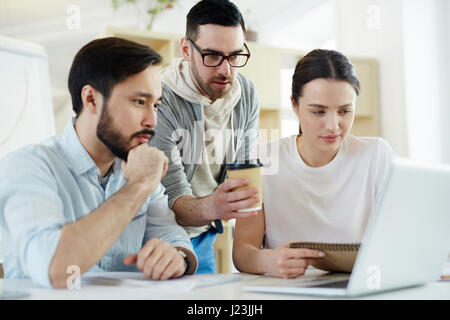 Group of focused business people looking at laptop screen while working in modern sunlit office Stock Photo