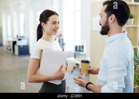 Portrait of smiling young woman talking to colleague during break in office, both holding paper coffee cups Stock Photo