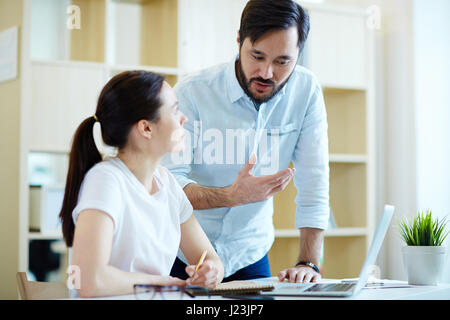 Portrait of adult Asian man leaning to help young woman at desk in office and discuss work Stock Photo