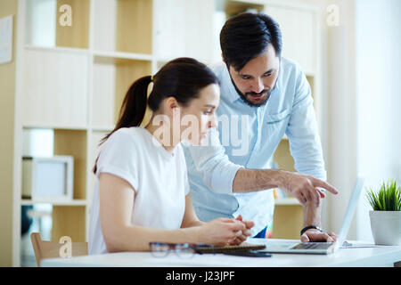 Portrait of adult Asian man leaning to help young woman at desk in office and discuss work, pointing at laptop Stock Photo