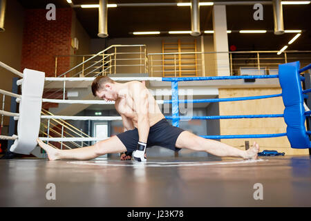 Portrait of muscular athlete stretching muscles in sports training, performing splits on floor in boxing ring Stock Photo