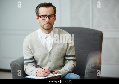 Serious psychiatrist in armchair making notes on paper Stock Photo