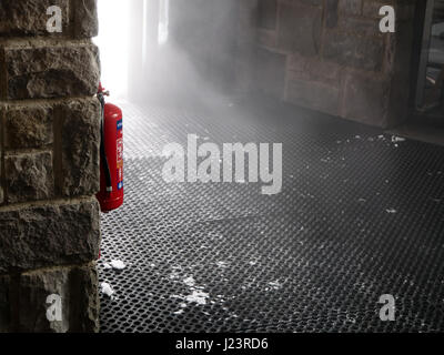 Fire extinguisher hang on the wall, cloud of mist look like smoke, comes through the door. Stock Photo