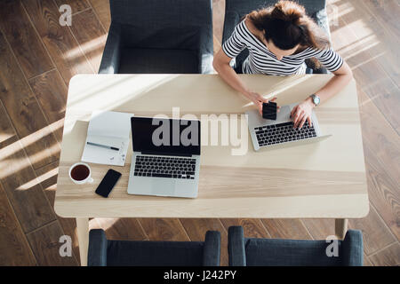 Top view of young woman sitting at desk and working on both her laptop and phone, while there's also another laptop and phone of her partner. Stock Photo