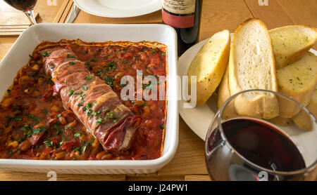 Close up Italian pork tenderloin on bed of tomatoes, beans and garlic with plate of garlic bread, glass of red wine Stock Photo
