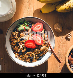 Delicious crunchy granola with nuts and raisins in bowl topped with fresh strawberries. Healthy breakfast food, morning sunlight, square crop. Stock Photo