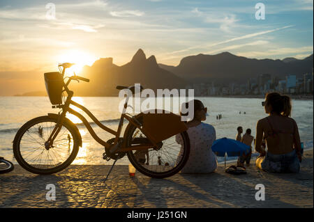 RIO DE JANEIRO - FEBRUARY 22, 2017: Friends gather next to a bicycle on the boardwalk to take in the sunset at Two Brothers Mountain. Stock Photo