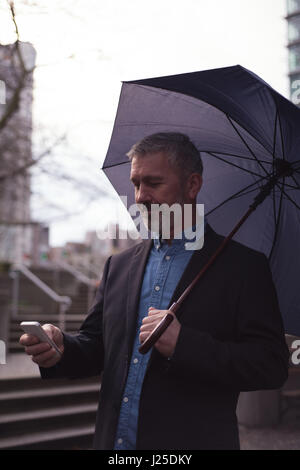 Businessman holding umbrella while using smart phone in city Stock Photo