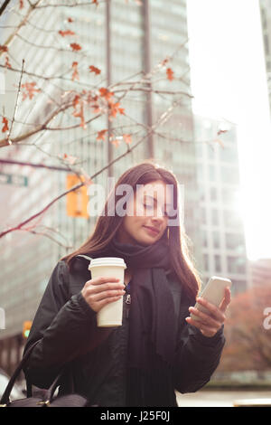 Woman holding coffee cup while using smartphone in city Stock Photo