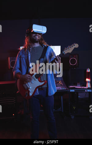 Male audio engineer using virtual reality headset while playing guitar in recording studio Stock Photo