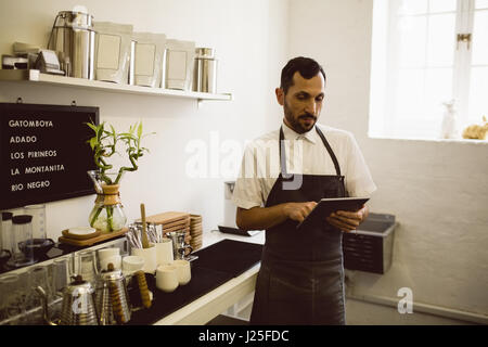 Barista using digital tablet while standing besides coffee grinding machine in coffee shop Stock Photo