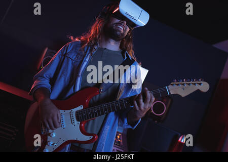 Male audio engineer using virtual reality headset while playing guitar in recording studio Stock Photo