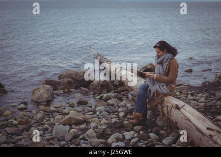 Woman using mobile phone while sitting on driftwood at beach Stock Photo