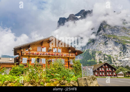 Traditional farmhouses in idyllic mountain scenery with high mountain peaks covered in clouds and fog on a beautiful sunny day in summer, Grindelwald Stock Photo