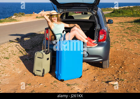 Travel, summer holidays and vacation concept - Young woman with suitcases on car trip. She is sitting in car back and gesture thumbs up. Stock Photo