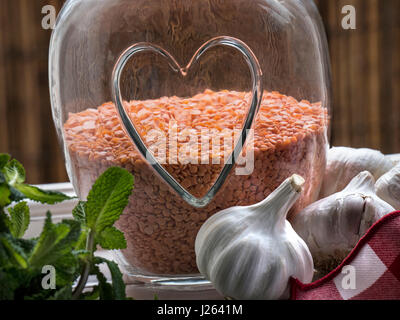 LENTILS GARLIC MINT Glass jar with healthy heart motif relief, a healthy edible pulse Legume, with garlics and sprig of mint by open kitchen window Stock Photo